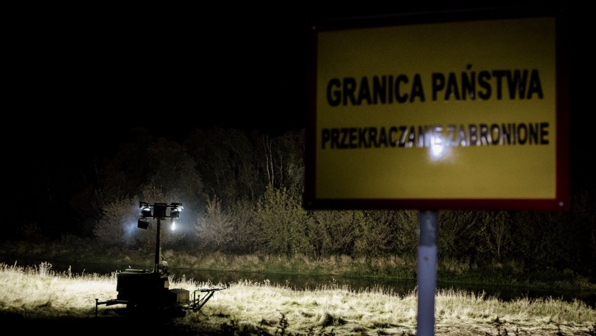 Jest noc, na pierwszym planie widoczna tabliczka "Granica państwa, przekraczanie zabronione". W oddali widać pracujący maszt oświetleniowy na zielonej granicy polsko-białoruskiej