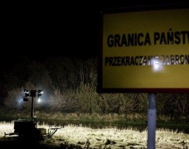 Jest noc, na pierwszym planie widoczna tabliczka "Granica państwa, przekraczanie zabronione". W oddali widać pracujący maszt oświetleniowy na zielonej granicy polsko-białoruskiej