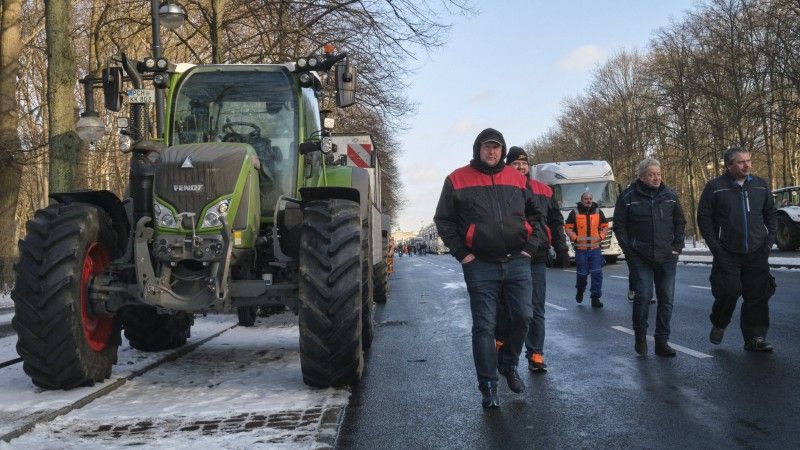 Rolnicy blokują ruch uliczny. Osoby idą po drodze, przy której zaparkowane są traktory.