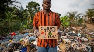 Chłopak trzyma tabliczkę z napisem „climate change is real”, stoi na tle wysypiska śmieci
