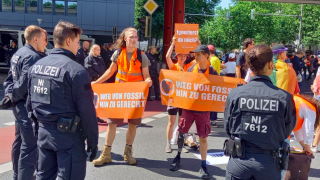 Aktywiści Ostatniego Pokolenia z pomarańczowymi transparentami protestują na ulicy, są otoczeni przez niemiecką policję