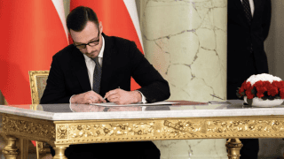 minister podpisuje dokument siedząc przy złoconym stole na tle flag Polski