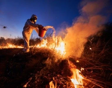 osoba w ochronnym kombinezonie gasi pożar traw wodą z wiadra