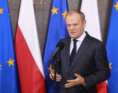 Premier Donald Tusk przemawia do mikrofonu na tle flag Polski i UE