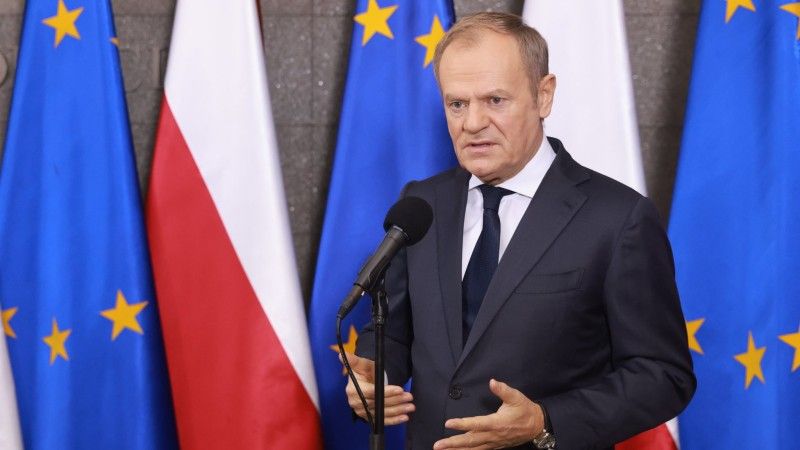 Premier Donald Tusk przemawia do mikrofonu na tle flag Polski i UE