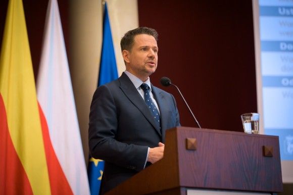 Rafał Trzaskowski za mównicą. W tle flagi Warszawy, Polski i UE
