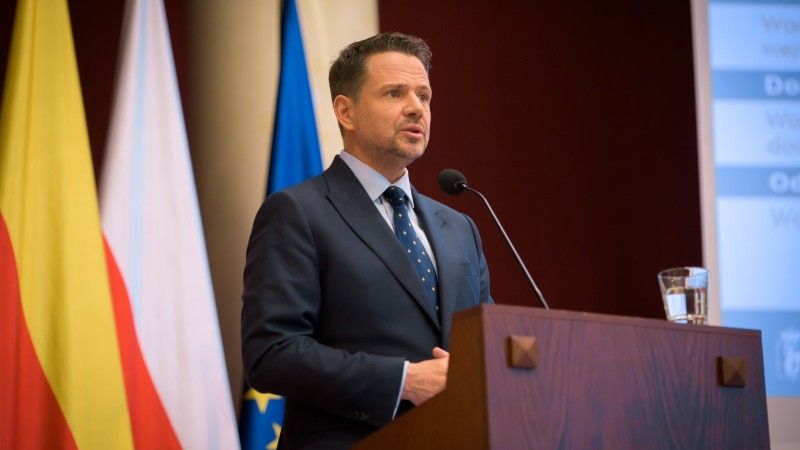 Rafał Trzaskowski za mównicą. W tle flagi Warszawy, Polski i UE