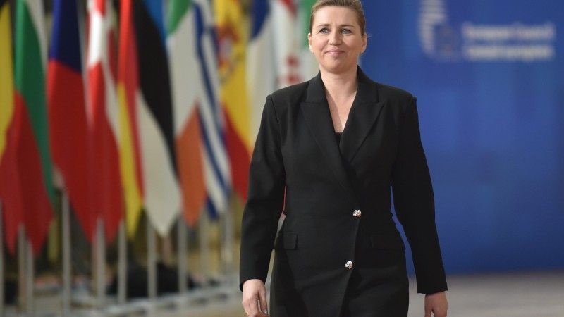 Mette Frederiksen idzie przez korytarz w budynku UE. W tle ścianka UE i flagi państw członkowskich