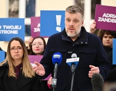 Zandber na wiecu partyjnym, przenawia do mikrofonów dziennikarzy. W tle osoby z partii Razem z transparentami: Adrian Zandberg, Po Twojej Stronie i logo partii
