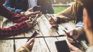 Osoby siedzące przy drewnianym stole, używające smartfonów, twarze niewidoczne