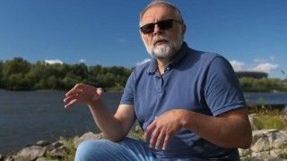 Profesor siedzi nad rzeką, opowiada o czymś gestykulując, w słoneczny ciepły dzień