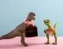 Dwa słodkie dinozaury na niebiesko różowym tle, mają na sobie hawajskie naszyjniki z różowych kwiatów, jeden trzyma walizkę