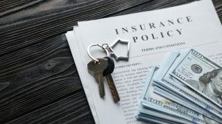 polisa ubezpieczeniowa na stole z ciemnego drewna, na której leży pęk dolarów oraz klucze z breloczkiem w kształcie domu