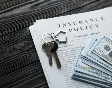 polisa ubezpieczeniowa na stole z ciemnego drewna, na której leży pęk dolarów oraz klucze z breloczkiem w kształcie domu