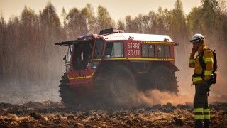 pojazd straży pożarnej na grubych wysokich oponach jedzie po torfowisku, obok strażak rozmawia przez krótkofalówkę