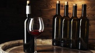 na drewnianej beczce na tle drewnianej ściany stoi 5 butelek wina i nalane czerwone wino do kieliszka