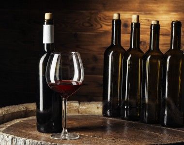 na drewnianej beczce na tle drewnianej ściany stoi 5 butelek wina i nalane czerwone wino do kieliszka