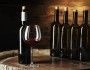 na drewnianej beczce na tle drewnianej ściany stoi 5 butelek wina i nalane czerwone wino do kieliszka