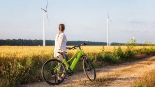 Kobieta stoi obok zielonego roweru elektrycznego, patrzy na turbiny wiatrowe w polu