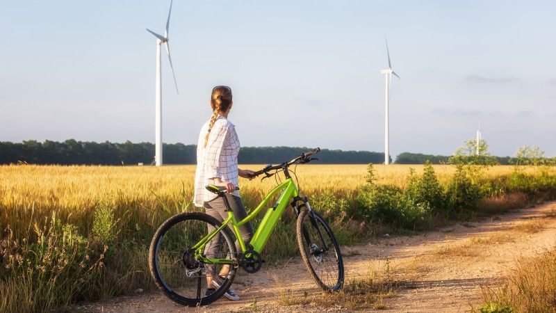 Kobieta stoi obok zielonego roweru elektrycznego, patrzy na turbiny wiatrowe w polu