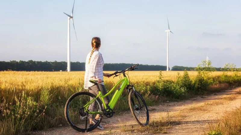 Kobieta stoi obok zielonego roweru elektrycznego, patrzy na turbiny wiatrowe w polu