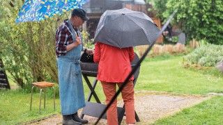 Starsza para grillująca w ogrodzie podczas deszczu z parasolkami w dłoniach