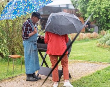 Starsza para grillująca w ogrodzie podczas deszczu z parasolkami w dłoniach