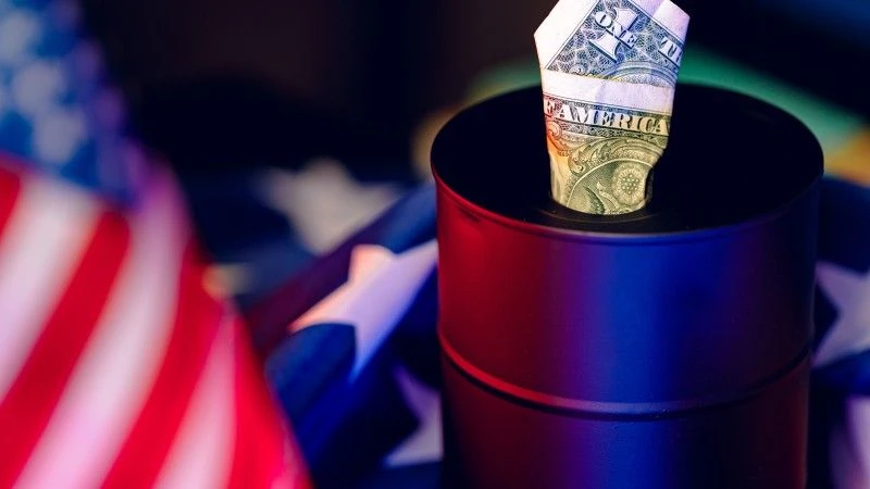 Baryłka ropy stoi na fladze USA. Z baryłki wystaje amerykański dolar