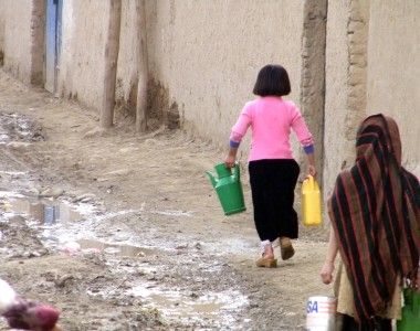 Dwie dziewczynki niosące kanistry z wodą po ulicy brudnej od błota – ulice w Kabulu bywają nieutwardzone. Jedna ma głowę nakrytą chustą, druga nie