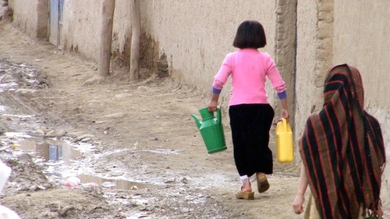 Dwie dziewczynki niosące kanistry z wodą po ulicy brudnej od błota – ulice w Kabulu bywają nieutwardzone. Jedna ma głowę nakrytą chustą, druga nie