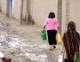 Dwie dziewczynki niosące kanistry z wodą po ulicy brudnej od błota – ulice w Kabulu bywają nieutwardzone. Jedna ma głowę nakrytą chustą, druga nie