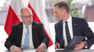 mężczyźni podpisują umowę siedząc za stołem, w tle flagi polski