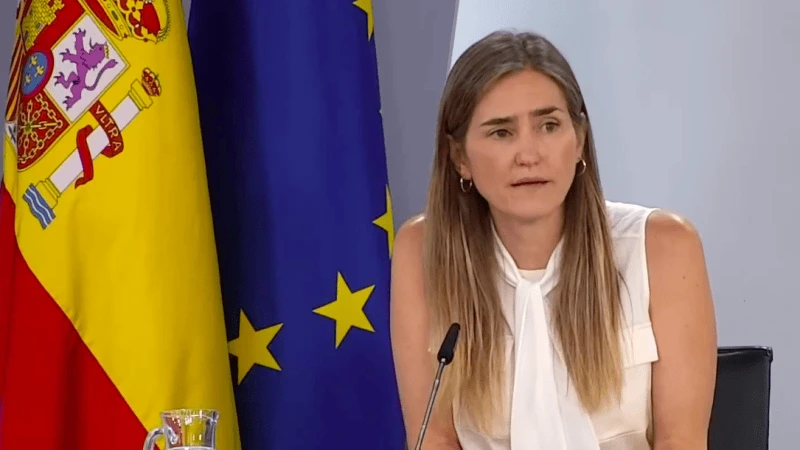 ministra przemawia na konferencji prasowej za stołem do mikrofonu, w tle flagi Hiszpanii i UE