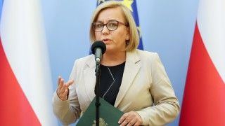 Paulina Hennig-Kloska przemawia do mikrofonu na tle flag Polski i UE, w ręku trzyma zieloną teczkę