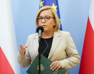 Paulina Hennig-Kloska przemawia do mikrofonu na tle flag Polski i UE, w ręku trzyma zieloną teczkę