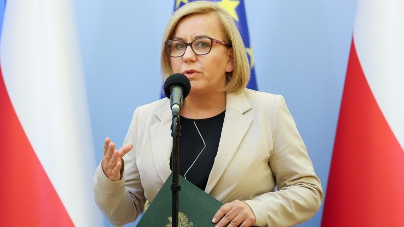 Paulina Hennig-Kloska przemawia do mikrofonu na tle flag Polski i UE, w ręku trzyma zieloną teczkę
