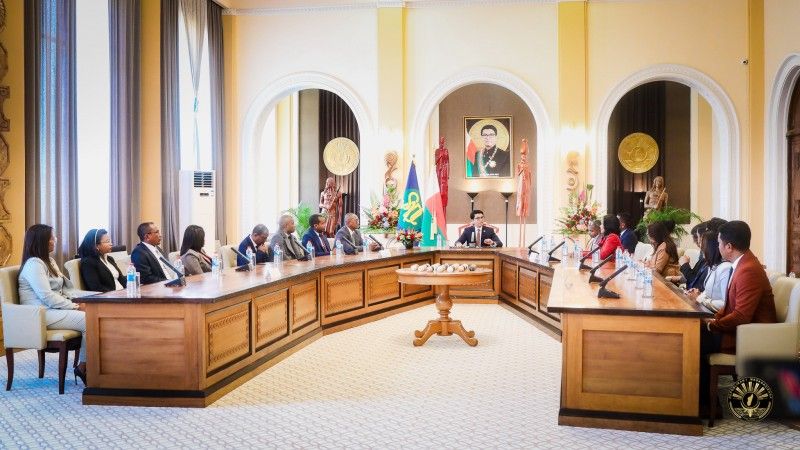 urzędnicy siedzą za stołem w kształcie podkowy, u szczytu siedzi prezydent, za nim wisi jego portret