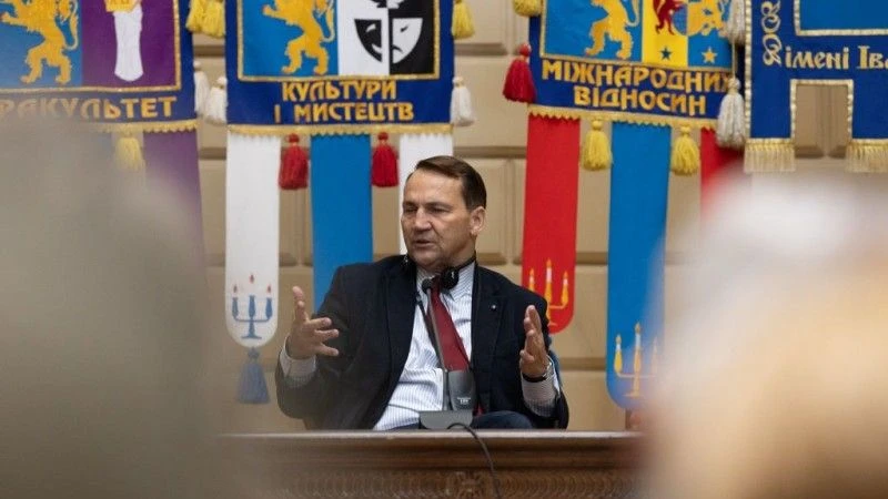 Man sits at a table with his hands up in front of several banners