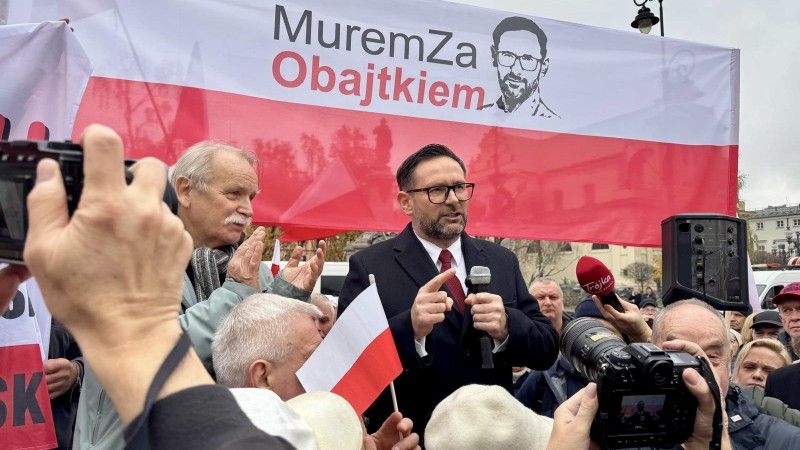 Obajtek przemawia do mikrofonu na podeście, wokół niego tłum ludzi, za nim flaga Polski z napisem: Murem za Obajtkiem oraz jego portretem (czarno-białym)
