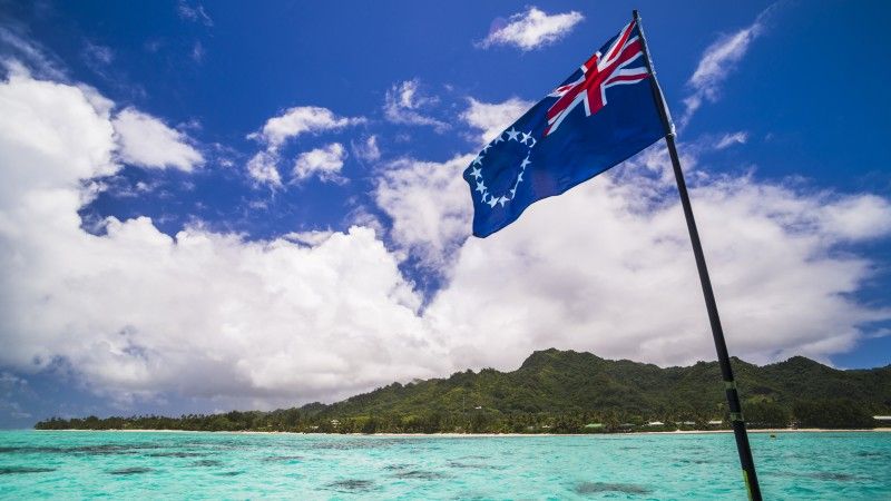 Flaga Wysp Cooka powiewa nad turkusową wodą przy małej wyspie z rajską plażą porośniętą lasem