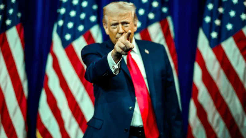 Prezydent stoi na tle flagi USA, wskazuje na coś przed sobą palcem