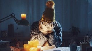 kobieta ubrana w sweter, czapkę i rękawiczki siedzi przy świeczkach przy biurku nad rachunkami, zrozpaczoną twarz ma ukrytą w dłoniach