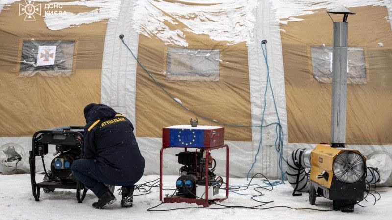 Pracownik służby obsługuje nagrzewnicę przy namiocie przysypanym śniegiem, obok na sprzęcie jest flaga Unii Europejskiej