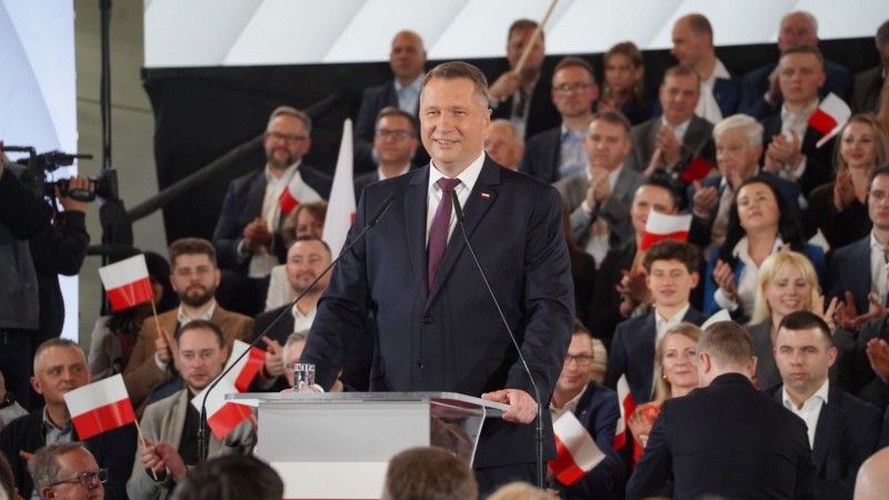 Czarnek stoi za mównicą, za nim na trybunach siedzą osoby z flagami Polski w rękach