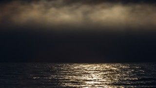 Wieczorne morze, odbija się w nim światło księżyce, nad nim dym lub chmury.