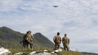 Four U.S. Navy SEAL operators in camouflage stand on a grassy Alaskan ridge with weathered wooden debris in the foreground, watching an AC-130J Ghostrider gunship fly overhead beneath a partly cloudy sky, with mountains in the background.