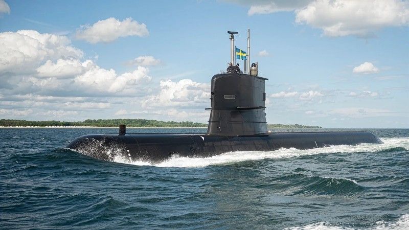 Swedish submarine HMS Södermanland sailing on the surface of the Baltic Sea, flying the Swedish naval ensign on its mast, with two crew members on the conning tower; a forested coastline and partly cloudy sky in the background.