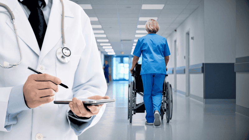 szpital: pielęgniarka z wózkiem inwalidzkim oraz lekarz z tabletem w dłoni.