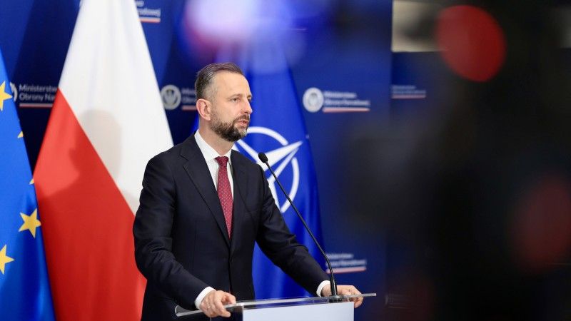 polityk przemawia zza mównicy na tle flag UE, Polski i NATO