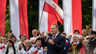 prezydent przemawia do mikrofonu na tle stojących ludzi i flag RP